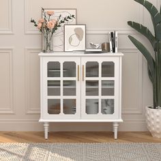 a white cabinet with glass doors next to a potted plant and pictures on the wall