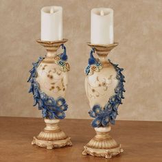 two white candles with blue flowers and leaves on them sitting next to each other in front of a wall