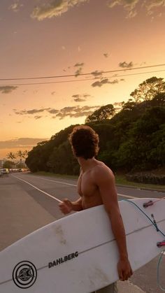 a shirtless man holding a surfboard on the side of the road at sunset