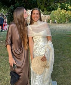 two women standing next to each other in front of trees and grass, one wearing a white dress