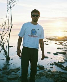 a man standing in front of the ocean with his hands on his hips wearing sunglasses