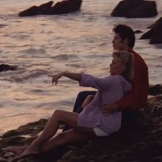 a man and woman are sitting on the rocks by the ocean pointing at something in the distance