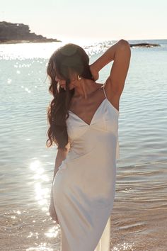 a woman in a white dress is standing on the beach with her hands behind her head