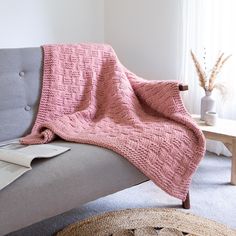 a pink knitted blanket sitting on top of a gray couch next to a window