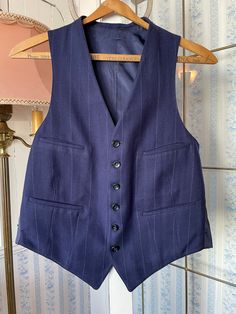 This tailored vest is made from a wool blend in dark royal/navy blue, with widely spaced light blue and beige pinstripes. It has the original buttons in the front and four front pockets, and is fully lined. There are no labels. The measurements, taken with the vest lying flat. are: shoulder to shoulder, 11 inches; armpit to armpit, 19 inches; length, 24 inches in front and 20 inches in back; bottom edge, 17 inches. In very good condition. Fitted Wool Vest For Tailoring, Classic Blue Three-piece Suit With Pockets, Classic Blue Three-piece Suit, Fitted Pinstripe Wool Outerwear, Pinstripe Vest For Work, Fitted Wool Sleeveless Blazer, Fitted Sleeveless Wool Blazer, Fitted Wool Vest With Welt Pockets, Tailored Pinstripe Sleeveless Vest