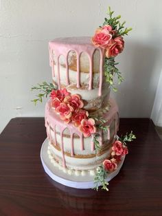 a three tiered cake with pink icing and flowers on the top, sitting on a wooden table