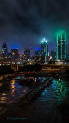 the city skyline is lit up in green and blue as rain falls on the ground