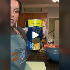 a woman holding up a can of beer in her kitchen while wearing an apron and looking at the camera