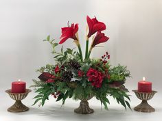 a vase filled with red flowers and greenery next to two candles on top of a table