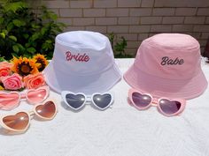 "Introducing our Bride Bucket Hat - a stylish and playful accessory designed to add a touch of fun and flair to the bride's special day! Whether you're lounging by the pool, enjoying a beachside celebration, or simply adding a playful element to your pre-wedding festivities, it is a must-have for any bride-to-be. Paired with the hat, our Heart Sunglasses complete the look with undeniable charm. The heart-shaped frames add a touch of whimsy and romance, creating a playful vibe that perfectly complements the bride's joyous spirit.  Bride Bucket Hat ✨Made of High-Quality 100% Cotton. ✨Comfortable And Durable. ✨Embroidered logo (Bride or Babe). ✨Colours (Available in White or Pink). ✨Relaxed fit, 22\" diameter (56-58cm). ✨Foldable (Easy storage in handbag or backpack)." White Novelty Mini Hats For Summer, Summer Wedding Mini Hat, Cute Pink Wedding Hat, White Adjustable Hat For Bachelorette Party, Adjustable Embroidered Party Hat, White Bucket Hat Mini Size, Bachelorette Bucket Hats, Custom White Bucket Hat, Playful White Bucket Hat