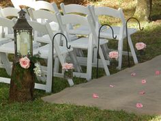 rows of white folding chairs with pink flowers on the grass near a tree trunk and walkway