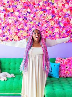 a woman with pink hair standing in front of a flower covered wall and green couch