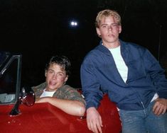 two young men sitting on the hood of a red car