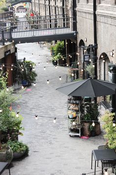 an outdoor cafe with tables and umbrellas on the sidewalk next to plants, potted trees and hanging lights