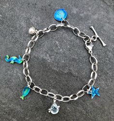 This silver charm bracelet is made from Sterling Silver and Enamel . The Silver Belcher chain which holds various enamel charms that capture the wonders of the ocean has  links that are each approx: 8mm long. It has an adjustable trigger so fits any size wrist. The charms are a starfish, shell, fish, sea horse and a Pearl and a tear-drop, faceted Aquamarine. The measurement is approximately: 8.5 inches but is adjustable. Enamel Bracelet Jewelry With Lobster Clasp, Enamel Bracelet With Lobster Clasp, Nickel-free Enamel Bracelet As A Gift, Blue Charm Bracelet With Dangling Charms As Gift, Enamel Charms Bracelets For Gift, Themed Bracelet With Lobster Clasp, Themed Jewelry With Lobster Clasp, Blue Metal Charm Bracelet With Lobster Clasp, Silver Enamel Jewelry With Dangling Charms