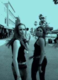 two young women standing next to each other on a city street with palm trees in the background