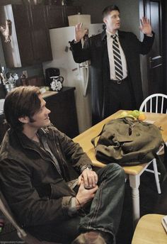 two men sitting at a table in a kitchen with one holding his hand up to the ceiling