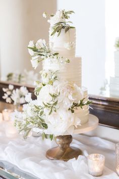 a wedding cake with white flowers and greenery sits on a table in front of candles