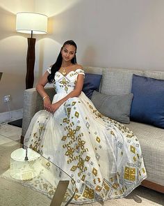 a woman is sitting on a couch in a white and gold dress with her legs crossed