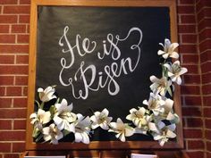 there is a sign that says he is risen with flowers in front of the chalkboard