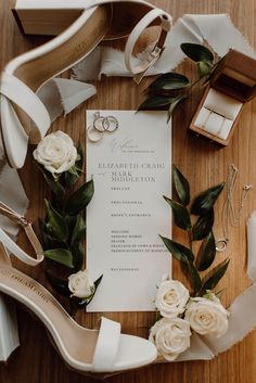 wedding stationery with white roses, rings and shoes on a table next to the bride's bouquet