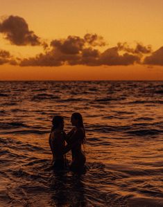 two people standing in the water at sunset with clouds above them and one person holding his arm around the other's neck