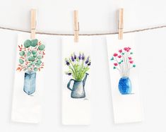 three watercolor paintings hanging on clothes pins with flowers in vases and blue vase