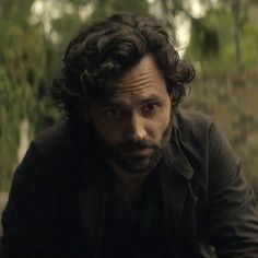 a man with curly hair and beard sitting in front of some trees looking at the camera