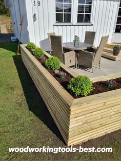 a wooden raised garden bed in front of a white building with patio furniture and flowers