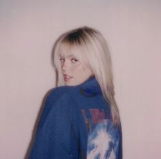 a woman with long blonde hair wearing a blue jacket and looking at the camera while standing in front of a white wall