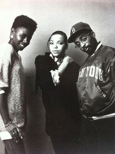 three people standing next to each other in front of a white wall and one person wearing a baseball cap