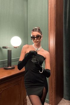 a woman in a black dress and gloves standing next to a desk with a light on it