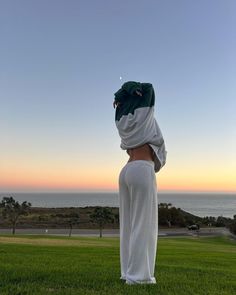 a woman in white pants standing on top of a lush green field next to the ocean