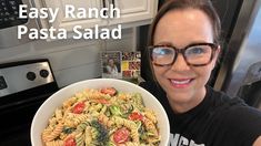 a woman holding a bowl of pasta with tomatoes and broccoli in it while smiling at the camera