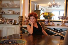 a woman sitting at a wooden table in front of a mirror and shelves with vases on it