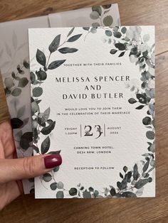 a hand holding up a wedding card with greenery on it and the wording reads,