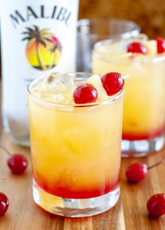 two glasses filled with drinks and cherries on top of a wooden table next to a bottle