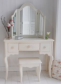 a white dressing table with a mirror, stool and flower vase on the top shelf