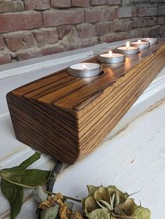 three candles are placed on a wooden shelf next to some flowers and leaves in front of a brick wall