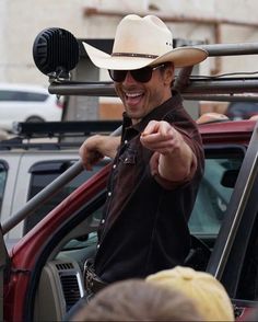 a man wearing a cowboy hat and sunglasses points at something in the distance while standing next to a red car