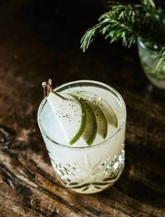 a glass filled with liquid and slices of apple on top of a wooden table next to a potted plant