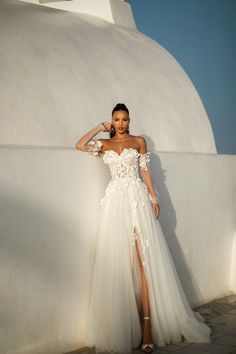 a woman in a white dress standing next to a wall and posing for the camera