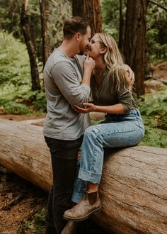 a man and woman sitting on a log in the woods, one is kissing the other's forehead