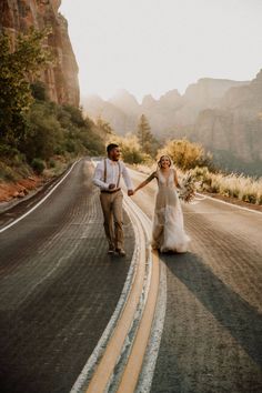 a bride and groom walking down the road holding hands in front of some mountains at sunset