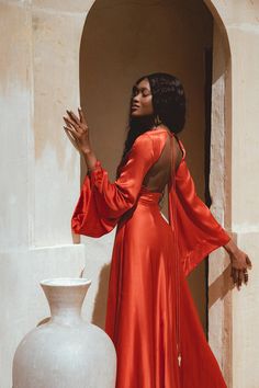 a woman in an orange dress standing next to a white vase and looking off into the distance