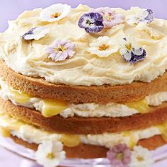 a layered cake with white frosting and purple flowers on top, sitting on a glass platter