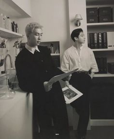 two men standing next to each other in front of a kitchen counter with books on it