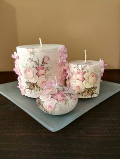 two white candles with pink flowers on them sitting on a blue plate next to a brown wall