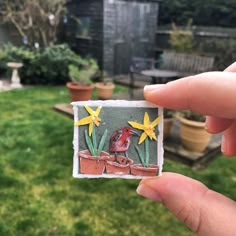 a hand holding up a small card with a bird and flowers on it in front of some potted plants