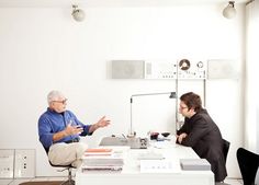 two men sitting at a table talking to each other in front of a white wall